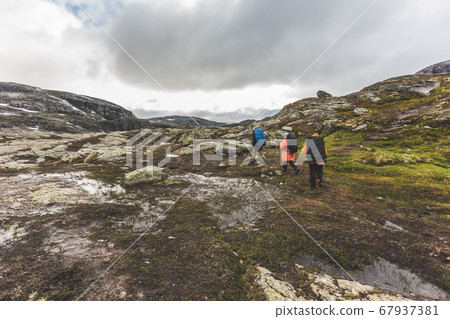 Tourists with backpacks walk across a rocky plain to the famous landmark - Troll tongue rock or Trolltunga. Cold and windy summer in Norway. Tourists with backpacks walk across a rocky plain to the famous landmark - Troll tongue rock or Trolltunga. Cold and windy summer in Norway. 67937381