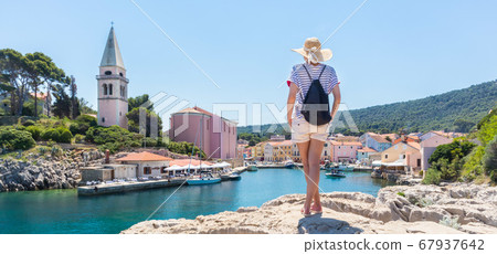 Woman traveler wearing straw summer hat and backpack, standing at edge of the rocky cliff enjoying beautiful panoramic view of Veli Losinj, Losinj island, Croatia 67937642
