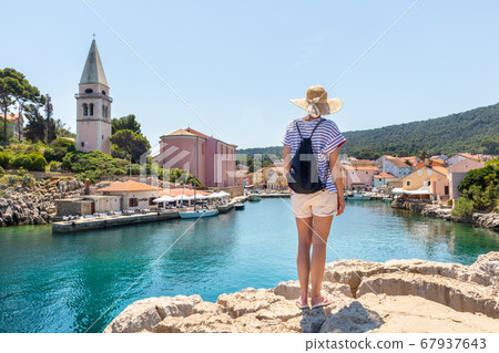 Woman traveler wearing straw summer hat and backpack, standing at edge of the rocky cliff enjoying beautiful panoramic view of Veli Losinj, Losinj island, Croatia 67937643