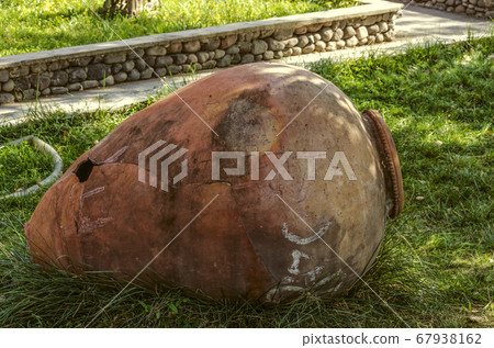 Large cracked pitcher of red clay is lying on the green grass in a rural courtyard 67938162