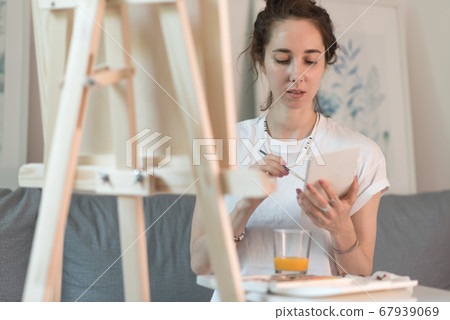 beautiful woman is sitting on sofa, holding paintbrush, colored polish, artist in white T-shirt. Easel table. At home in workshop. Emotions of comfort, pleasure, inspiration creativity. Draws picture. 67939069