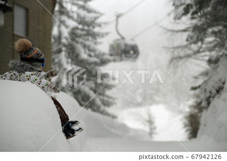 Small child in ski mask sitting on big snowbank under snowfall. Background of snowy trees, ski cabin going up on cabel Small child in ski mask sitting on big snowbank under snowfall. Background of snowy trees, ski cabin going up on cabel 67942126