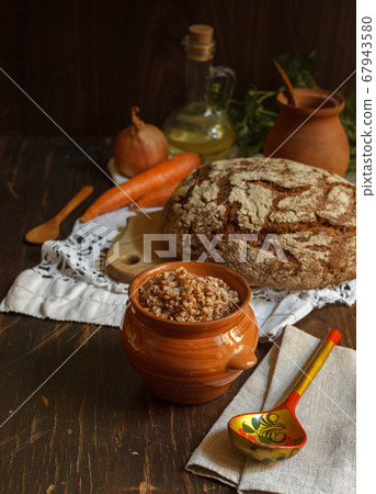 Buckwheat porridge in a pot and a wooden painted spoon on a linen napkin, black bread and vegetables in the background Buckwheat porridge in a pot and a wooden painted spoon on a linen napkin, black bread and vegetables in the background 67943580