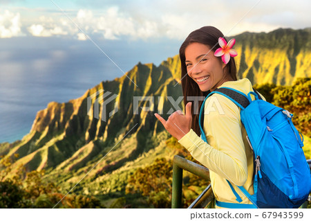 Hawaii hiker girl tourist doing shaka hawaiian hand sign at Na pali lookout in Kauai, Hawaii. Travel Asian woman backpacker hiking with bag at famous destination Kalalau hawaiian attraction Hawaii hiker girl tourist doing shaka hawaiian hand sign at Na pali lookout in Kauai, Hawaii. Travel Asian woman backpacker hiking with bag at famous destination Kalalau hawaiian attraction 67943599