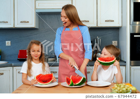 Sweet family, mother and her kids eating watermelon in their kitchen having fun Sweet family, mother and her kids eating watermelon in their kitchen having fun 67944472