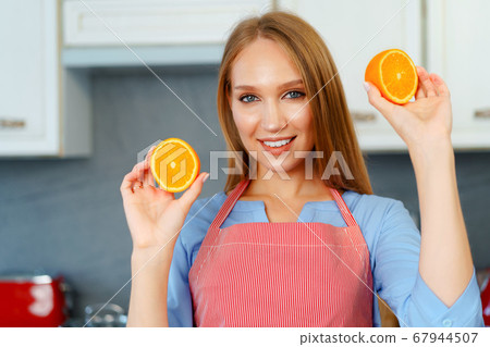 Beautiful caucasian woman in red apron holding ripe oranges while standing in kitchen Beautiful caucasian woman in red apron holding ripe oranges while standing in kitchen 67944507