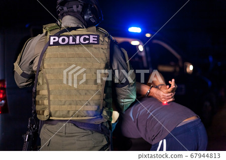 A police officer puts handcuffs on a criminal's hands during an arrest. Police car with flashing lighthouses. Blurried A police officer puts handcuffs on a criminal's hands during an arrest. Police car with flashing lighthouses. Blurried 67944813