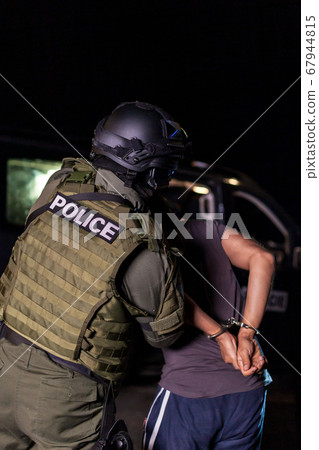 A police officer puts handcuffs on a criminal's hands during an arrest. Police car with flashing lighthouses. Blurried A police officer puts handcuffs on a criminal's hands during an arrest. Police car with flashing lighthouses. Blurried 67944815