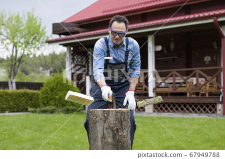 Adult man in jeans shirt and overalls chopping wood with axe in rural Adult man in jeans shirt and overalls chopping wood with axe in rural 67949788