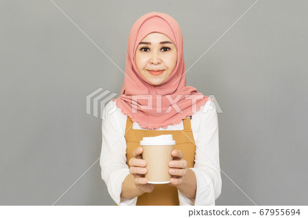 Small business owner happy young beautiful asian muslim woman propose hand arm paper cups hot beverage invite visit cafe wear traditional dress and brown apron isolated gray background 67955694