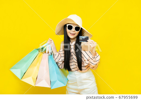 Beautiful excited happy young shopaholic asian woman wearing sungalsses and floppy hat posing isolated over yellow background holding pastel shopping bags ,summer sale concept 67955699