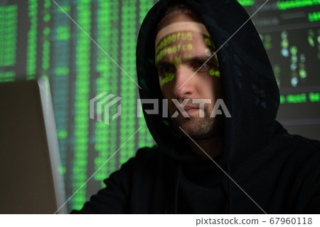 Male hacker working on a computer while green code characters reflect on his face in a dark office room. Male hacker working on a computer while green code characters reflect on his face in a dark office room. 67960118