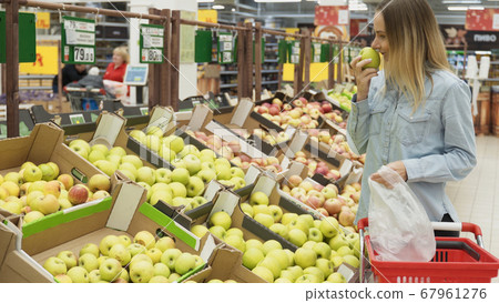 Portrait of a smiling attractive young woman shopper in the grocery shop during choosing and buying fresh apples at fruit department Portrait of a smiling attractive young woman shopper in the grocery shop during choosing and buying fresh apples at fruit department 67961276