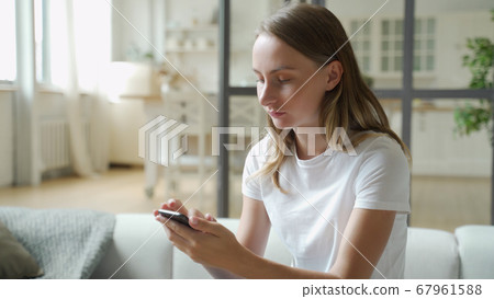 Young smiling woman using mobile phone while sitting on sofa at home 67961588