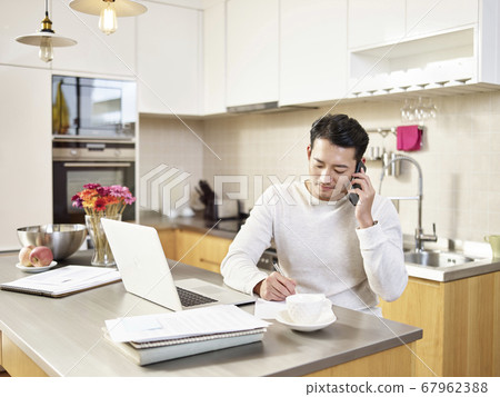 young asian business man sitting kitchen counter working at home talking on cellphone young asian business man sitting kitchen counter working at home talking on cellphone 67962388