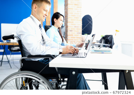 Businessman in wheelchair with colleague in office at working table Businessman in wheelchair with colleague in office at working table 67972324