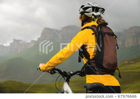 close-up young slender girl with a backpack and a bicycle helmet sits on a bicycle high in the mountains on a cloudy day and looks at the mountains and clouds. 67976666