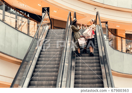 Two girls on an escalator in a shopping center. In the hands of paper bags. Shopping, leisure and entertainment. Two girls on an escalator in a shopping center. In the hands of paper bags. Shopping, leisure and entertainment. 67980363