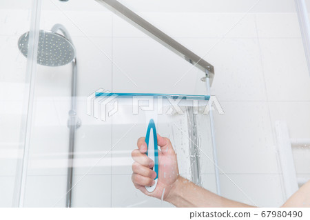 hand with glass scraper cleans glass in shower 67980490