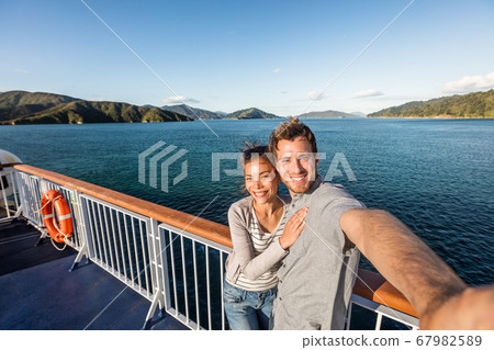 Cruise couple tourists taking selfie on New Zealand travel. People traveling on ferry boat Marlborough sounds taking self-portrait picture with mobile phone smiling at camera. Cruise couple tourists taking selfie on New Zealand travel. People traveling on ferry boat Marlborough sounds taking self-portrait picture with mobile phone smiling at camera. 67982589