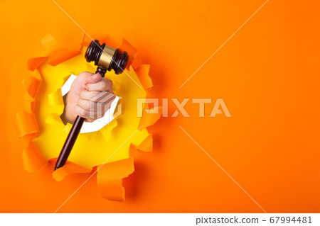 Hand holding a judge's gavel through torn orange 67994481