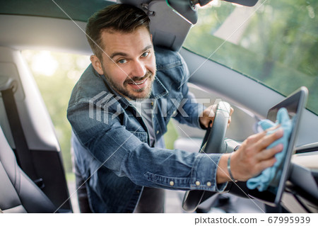 Close up portrait of smiling handsome Caucasian man with beard wiping dust from touchscreen display with microfiber cloth, inside his modern self-steering car and looking at camera Close up portrait of smiling handsome Caucasian man with beard wiping dust from touchscreen display with microfiber cloth, inside his modern self-steering car and looking at camera 67995939
