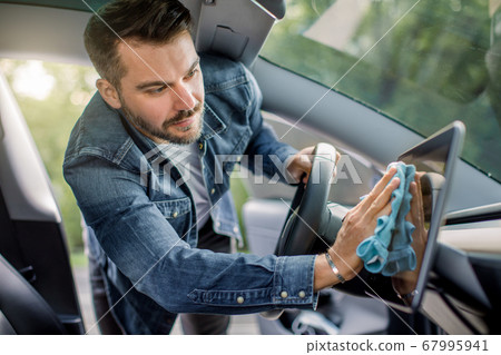 Young handsome Caucasian man cleaning the car console and touchscreen display of his modern electric car with microfiber cloth. Car wash and detailing concept at self car wash station Young handsome Caucasian man cleaning the car console and touchscreen display of his modern electric car with microfiber cloth. Car wash and detailing concept at self car wash station 67995941