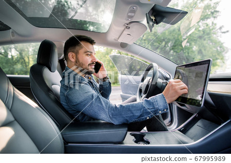 Testing a new electric futuristic car with self driving system. Side view of satisfied Caucasian man in casual jeans shirt sitting in modern car, talking phone and toushing navigation autopilot screen Testing a new electric futuristic car with self driving system. Side view of satisfied Caucasian man in casual jeans shirt sitting in modern car, talking phone and toushing navigation autopilot screen 67995989