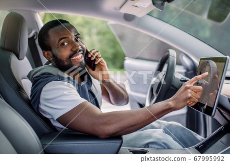 Smiling African American young guy talking phone while sitting in the cabin of a comfortable modern electric car and touching the screen of navigation self-driving autopilot. Testing a new car 67995992