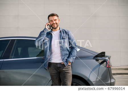 Smiling Caucasian man in casual wear talking phone while leaning on his luxury new electric car, standing on the charging station for charging a car. Electric car charging concept Smiling Caucasian man in casual wear talking phone while leaning on his luxury new electric car, standing on the charging station for charging a car. Electric car charging concept 67996014
