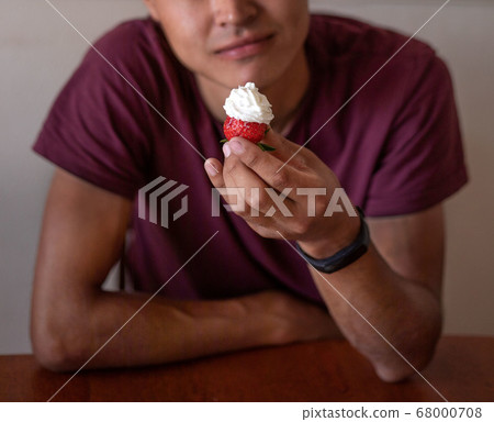 Guy is sitting at the table and holding strawberry with cream in his hand. Delicious food, dessert 68000708