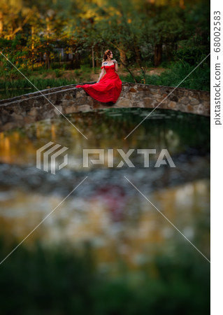 Redhead woman in red dress sitting on stone bridge Redhead woman in red dress sitting on stone bridge 68002583