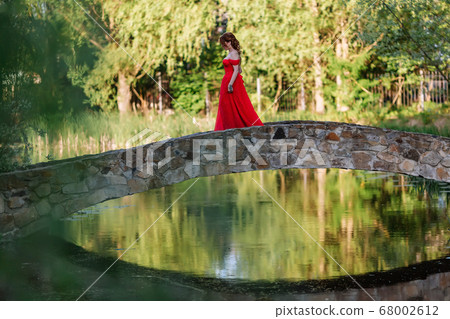 Redhead woman in red dress walking on stone bridge Redhead woman in red dress walking on stone bridge 68002612