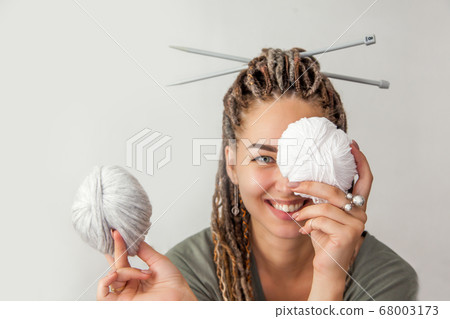 A beautiful young girl knitter smiles and holds skeins of light gray and white yarn in her hands. Woman with dreadlocks. A beautiful young girl knitter smiles and holds skeins of light gray and white yarn in her hands. Woman with dreadlocks. 68003173
