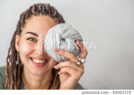 A beautiful young girl knitter smiles and holds skeins of light gray and white yarn in her hands. Woman with dreadlocks. A beautiful young girl knitter smiles and holds skeins of light gray and white yarn in her hands. Woman with dreadlocks. 68003179