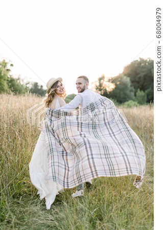 Young pregnant couple in the field during summer sunset. Handsome boy and his pretty pregnant girl hugging each other, covered with checkered blanket in the evening sun glow 68004979