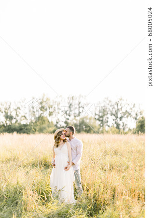 Young beautiful pregnant couple in white clothes walking in a field in the summer at sunset. Handsome bearded young man kissing his pretty pregnant lady in white dress and straw hat Young beautiful pregnant couple in white clothes walking in a field in the summer at sunset. Handsome bearded young man kissing his pretty pregnant lady in white dress and straw hat 68005024