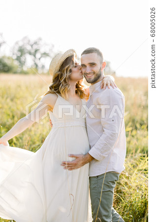 Waiting for the baby, pregnancy concept. Happy young couple, expecting a baby, walking in bright summer sunny day on a field. Man hugs and kisses his pretty pregnant woman in hat and dress Waiting for the baby, pregnancy concept. Happy young couple, expecting a baby, walking in bright summer sunny day on a field. Man hugs and kisses his pretty pregnant woman in hat and dress 68005026