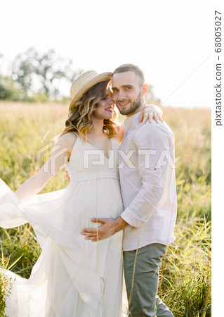 Waiting for the baby, pregnancy concept. Happy young couple, expecting a baby, walking in bright summer sunny day on a field. Man hugs and kisses his pretty pregnant woman in hat and dress Waiting for the baby, pregnancy concept. Happy young couple, expecting a baby, walking in bright summer sunny day on a field. Man hugs and kisses his pretty pregnant woman in hat and dress 68005027