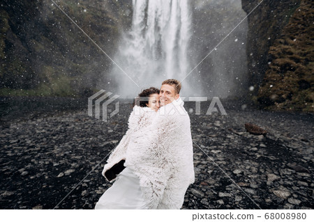 Wedding couple near Skogafoss waterfall. The bride and groom covered with a woolen blanket, where hugging. Snow is falling, falling snowflakes. 68008980