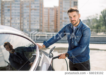 Horizontal shot of young Caucasian man looking at camera while charging his luxury electric car at outdoor station in front of modern new city buildings Horizontal shot of young Caucasian man looking at camera while charging his luxury electric car at outdoor station in front of modern new city buildings 68019102