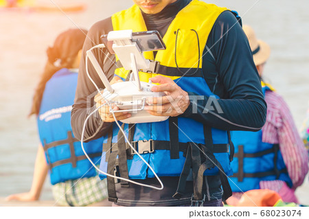 Young man in blue and yellow life vest controlling a drone to take pictures of ocean coast with tourists while traveling on a raft in the sea. Hands holding drone remote controller. Young man in blue and yellow life vest controlling a drone to take pictures of ocean coast with tourists while traveling on a raft in the sea. Hands holding drone remote controller. 68023074