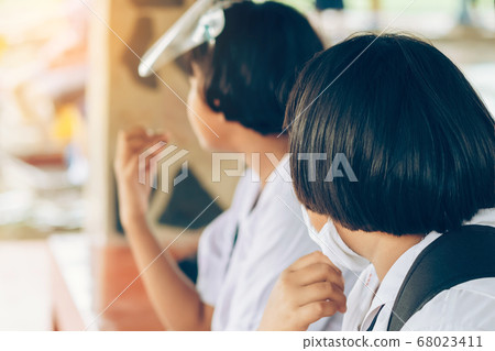Female elementary school student wear face mask to prevent the Coronavirus(Covid-19) wait for her parents to pick her up to return home after school and the rain just stop in front of the school gate 68023411