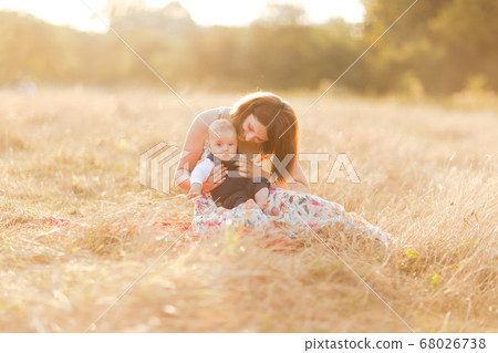 Family with children walking outdoors in summer field at sunset. Pretty young mother and her little baby boy sitting on the grass, having fun in summer field. People, family day and lifestyle concept Family with children walking outdoors in summer field at sunset. Pretty young mother and her little baby boy sitting on the grass, having fun in summer field. People, family day and lifestyle concept 68026738