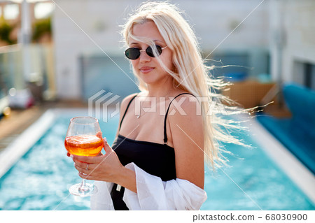Attractive young woman wearing bikini, sunglasses, standing on the swimming pool, drinking cocktails and dancing Attractive young woman wearing bikini, sunglasses, standing on the swimming pool, drinking cocktails and dancing 68030900
