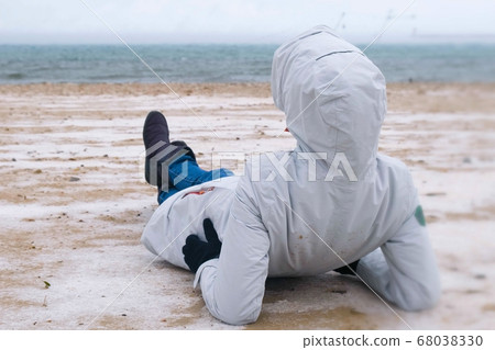 Woman tourist lies on a snowy sandy beach in winter by the sea. Woman in a jacket and jeans, back view. 68038330