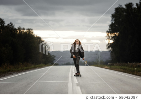 Man in office suit is riding skateboard longboard down road outside the city. 68042087