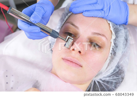 Cosmetologist makes a micro-current procedure for the face of a young woman. Face close-up, front view. Cosmetologist makes a micro-current procedure for the face of a young woman. Face close-up, front view. 68043349
