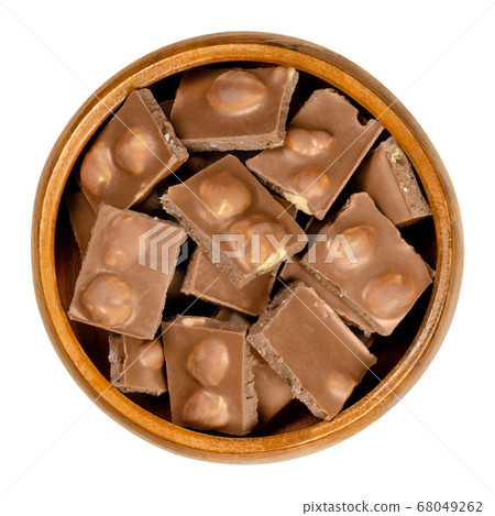 Hazelnut milk chocolate, bars broken into pieces, in wooden bowl. Small segments of candy bar. Milk chocolate with whole roasted hazelnuts, ready-to-eat. Sweet dessert. Close-up from above food photo. 68049262