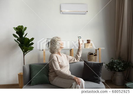 Elderly woman holding airconditioner remote control set comfortable temperature indoors Elderly woman holding airconditioner remote control set comfortable temperature indoors 68052925